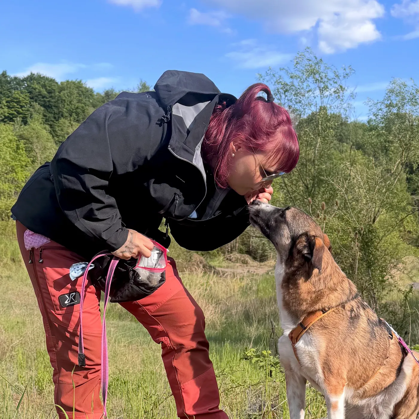 Eine Frau mit pinken Haaren beugt sich Nase an Nase zu ihrem sitzenden Hund auf einer Wiese