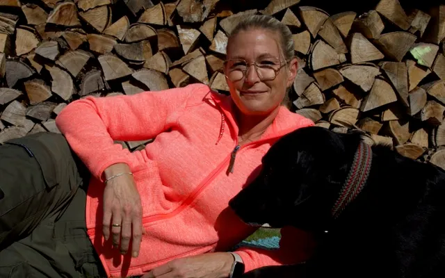 Eine Frau lehnt an einem Stapel Brennholz, neben ihr liegt ein schwarzer Hund auf einer blauen Decke